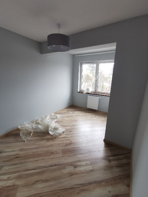 Pokój po świeżym malowaniu na szaro, z ułożonymi panelami podłogowymi, zabezpieczonymi folią ochronną w rogu, widoczne okno z grzejnikiem.