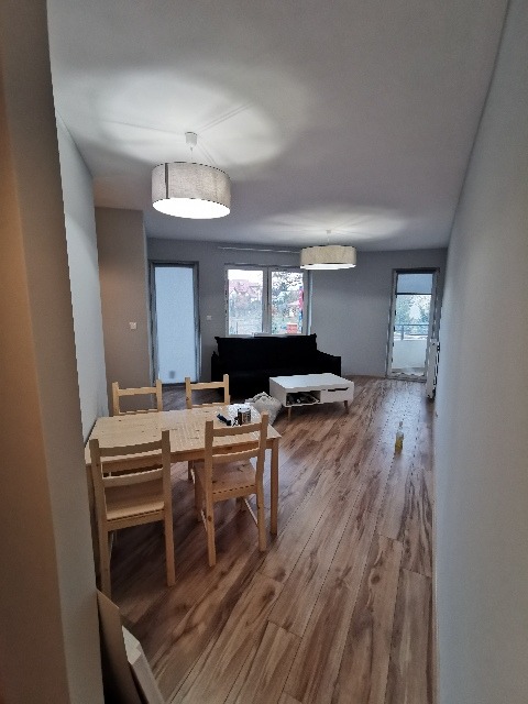 Jasne, przestronne wnętrze po remoncie w Warszawie, z drewnianą podłogą panelową, widoczny stół z krzesłami i kanapa w tle. Na podłodze widoczne ślady po remoncie.