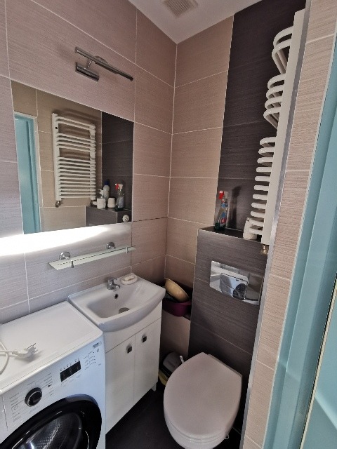 Ukończona łazienka z umywalką na szafce, pralką, toaletą, grzejnikiem drabinkowym i lustrem oświetlonym halogenem, wykończona płytkami w odcieniach beżu i szarości.