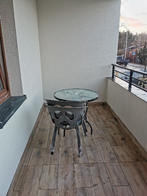 Balkon z drewnopodobnymi płytkami, metalowym stolikiem z szklanym blatem i ażurowymi krzesłami, widok na ulicę.