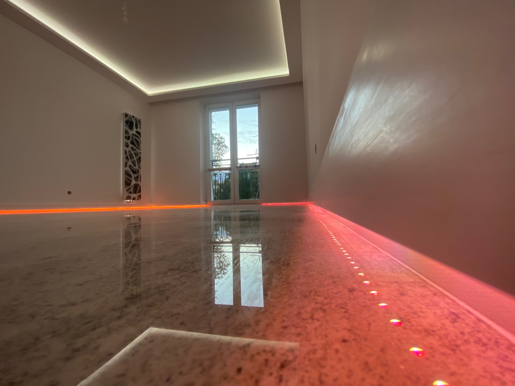 Błyszcząca, jasna posadzka z kamienia naturalnego z podświetleniem LED wzdłuż ściany i sufitu, odbijająca okno balkonowe i dekoracyjny panel ścienny.