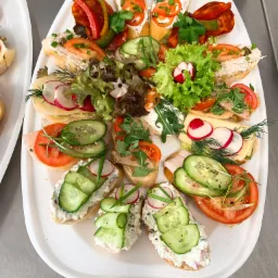 Elegancko podane, różnorodne kanapki na białej tacy cateringowej: z ogórkiem i twarożkiem, wędliną, serem, pomidorami, rzodkiewką i papryką, udekorowane zieleniną.