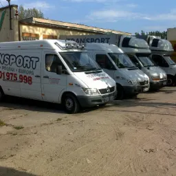 Flota białych samochodów dostawczych z napisem TRANSPORT i numerem telefonu, zaparkowana przed budynkiem firmy w Kołobrzegu. Pierwszy samochód z widocznym napisem 'Kołobrzeg Polska Europa'.