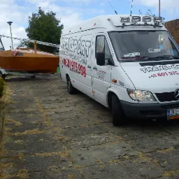 Biały bus transportowy z napisem 'TRANSPORT Kołobrzeg Polska Europa' i numerem telefonu, holuje na przyczepie małą łódź żaglową.