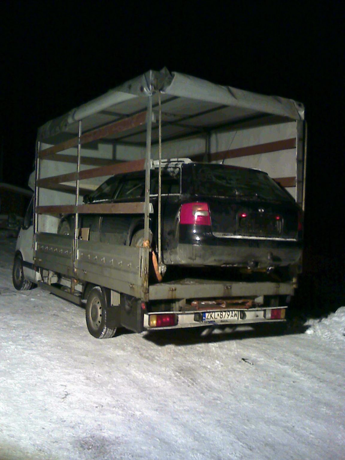 Czarny samochód osobowy typu kombi zabezpieczony pasami transportowymi na platformie niskopodwoziowej pojazdu ciężarowego z plandeką w zimowych warunkach nocnych. Widoczna tablica rejestracyjna ZKL...