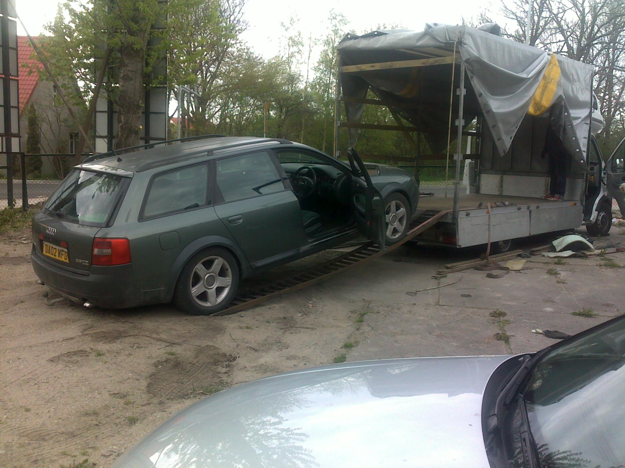 Zielone kombi Audi z otwartymi drzwiami wjeżdża po rampie na platformę transportową przykrytą szarą plandeką, widoczny numer rejestracyjny z Wielkiej Brytanii.