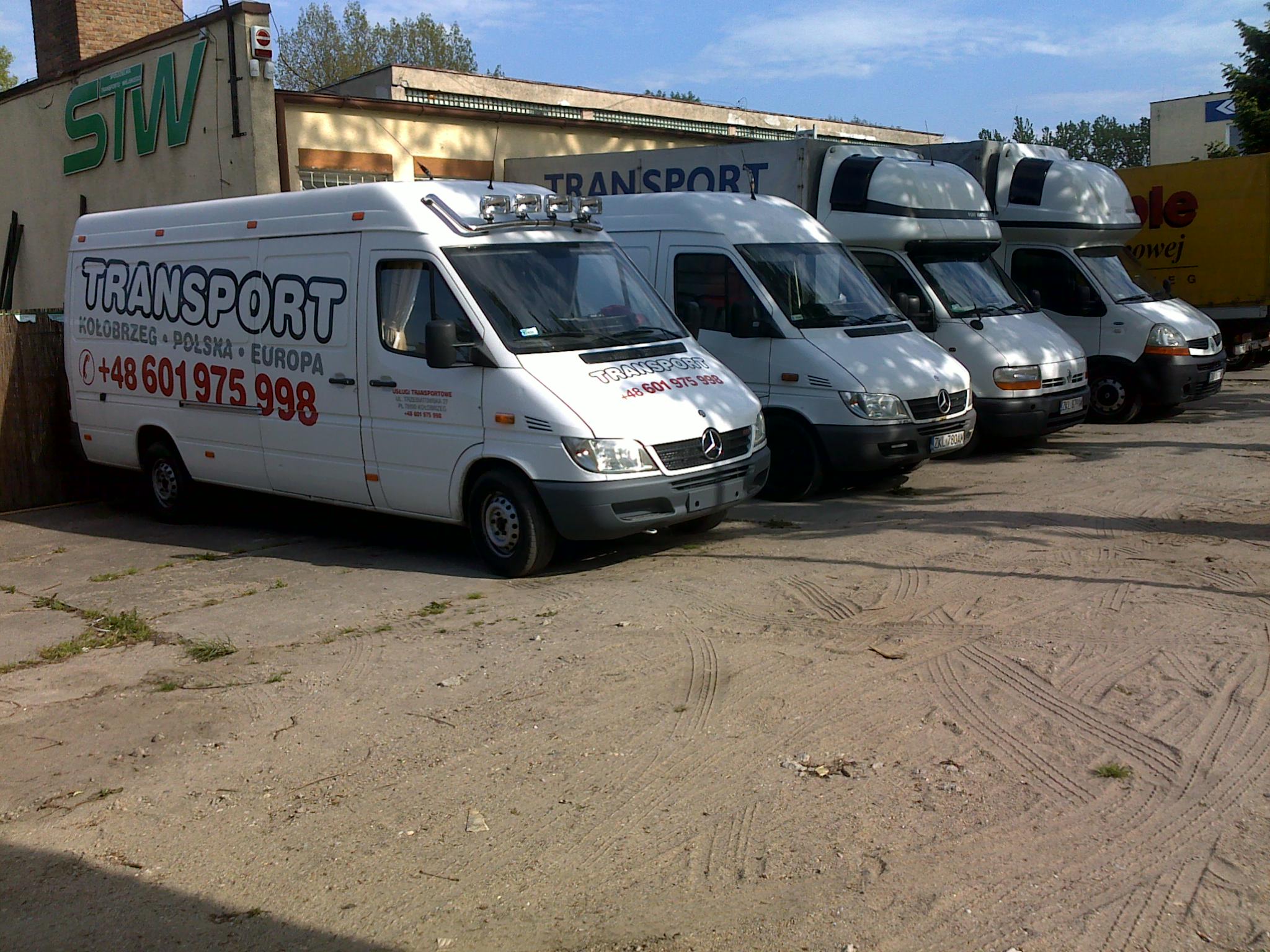 Rząd białych samochodów dostawczych Mercedes-Benz Sprinter i Renault Master z zabudową kontenerową, zaparkowanych na betonowym placu przed budynkiem STW, z napisem 'TRANSPORT Kołobrzeg Polska...