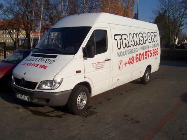 Biały bus transportowy z napisem 'TRANSPORT Kołobrzeg Polska Europa +48 601 975 998' zaparkowany na brukowanym placu, widoczny z boku, z budynkiem w tle.