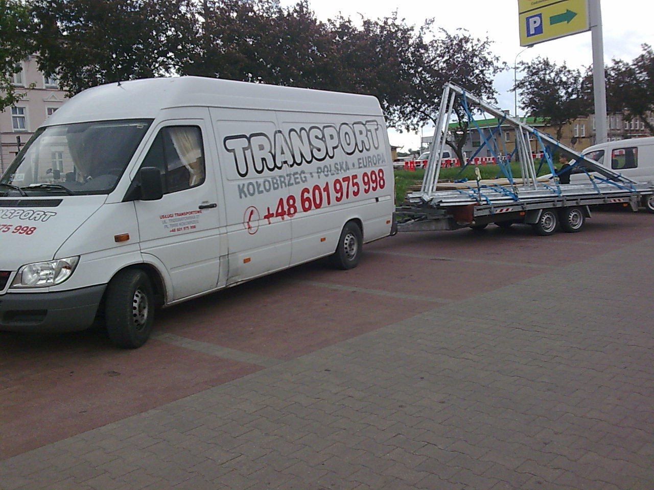 Biały bus z napisem 'TRANSPORT KOŁOBRZEG POLSKA EUROPA' ciągnie przyczepę z metalową konstrukcją, zaparkowany na placu z kostki brukowej.
