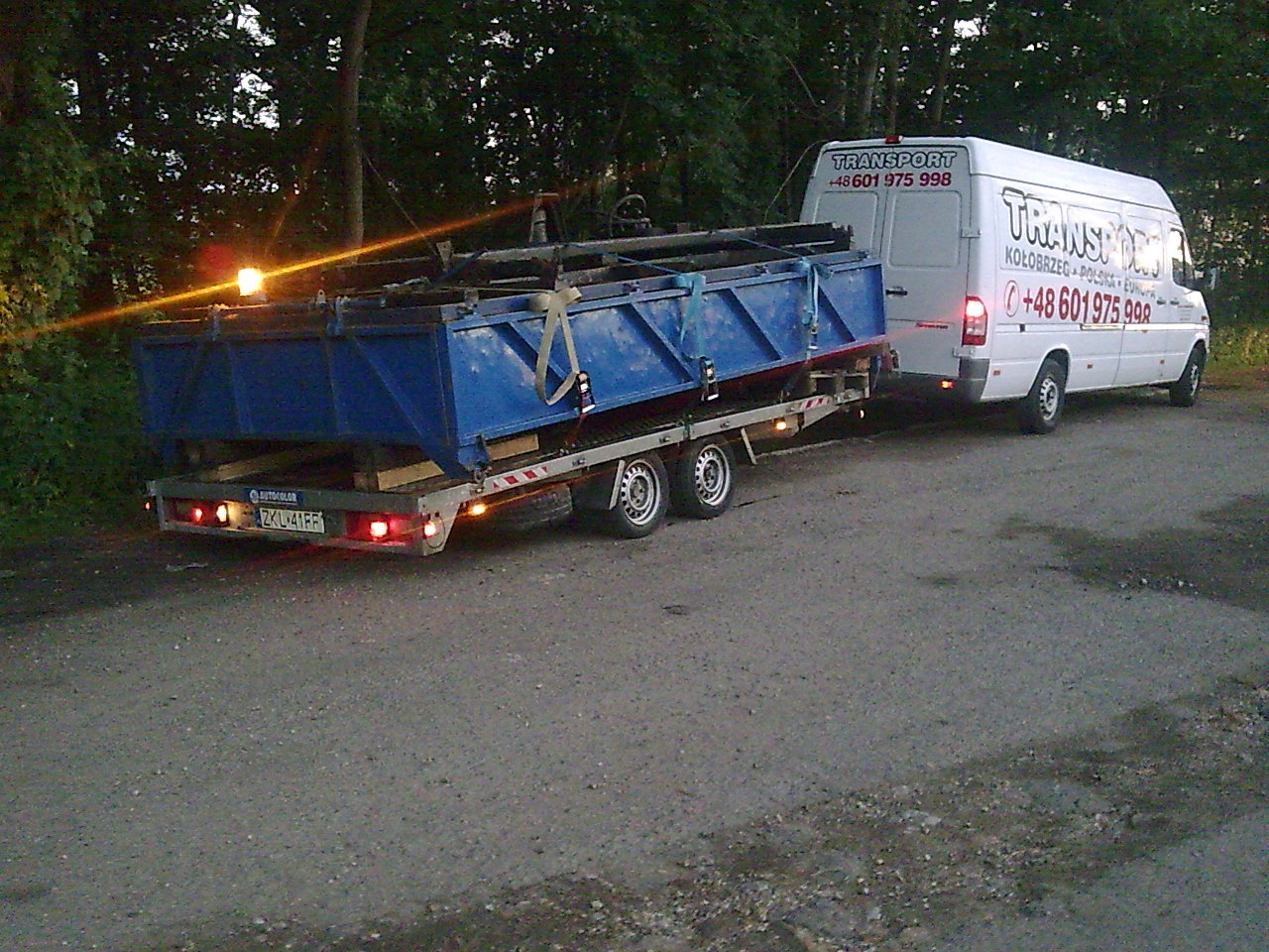 Laweta z niebieskim kontenerem transportowana busem z napisem TRANSPORT Kołobrzeg Polska Europa, widok z tyłu, oświetlenie wieczorne.