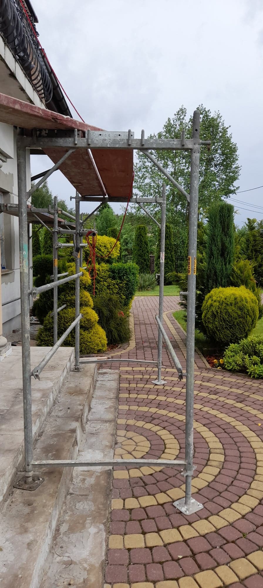 Metalowe rusztowanie ustawione przy schodach budynku, z widoczną czerwoną liną i ozdobnym brukowanym chodnikiem w tle.