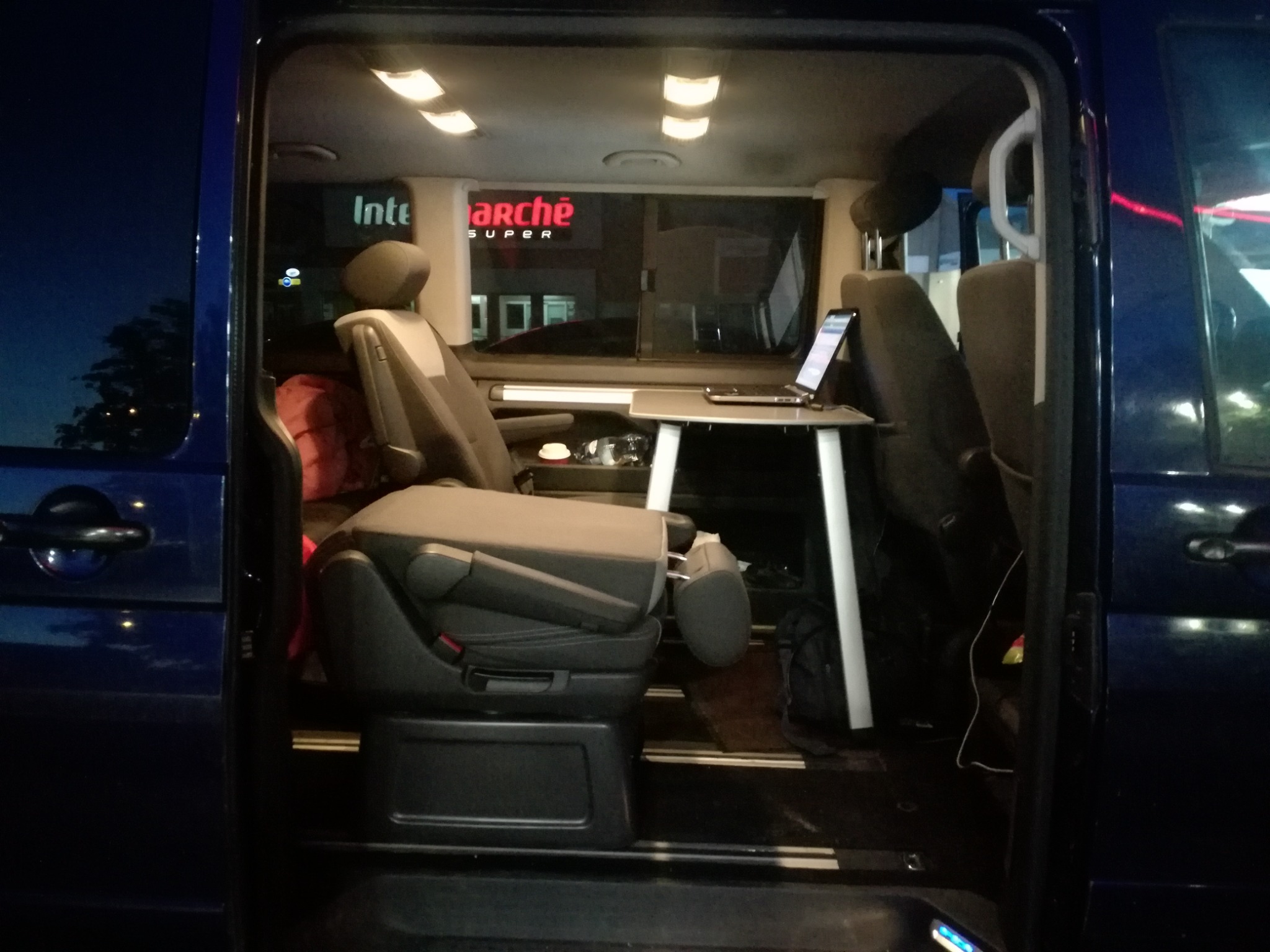 Wnętrze niebieskiego vana z rozłożonym stolikiem, laptopem i telefonem, widok z otwartych drzwi, w tle oświetlony budynek sklepu.