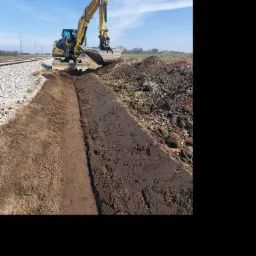 Żółta koparka Yanmar w trakcie wykonywania wykopu rowu melioracyjnego wzdłuż torów kolejowych, na tle błękitnego nieba z delikatnymi obłokami.