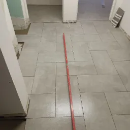 Układanie szarych płytek podłogowych w korytarzu z użyciem poziomicy i krzyżyków dystansowych, widoczne niedokończone fragmenty przy futrynach.