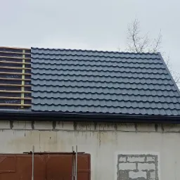 Częściowo pokryty dach blachodachówką w kolorze grafitowym, widoczne drewniane łaty i membrana, rynna, fragment elewacji budynku z cegły silikatowej i metalowe schody.