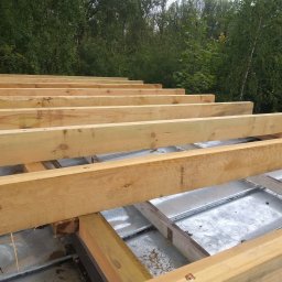 Tomasz Groblewski - Drewniane belki konstrukcyjne układane na metalowym poszyciu dachu, widok z góry na tle zieleni drzew.