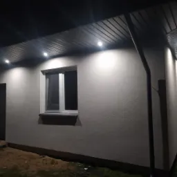 Oświetlona elewacja budynku jednorodzinnego w nocy, widoczne podbitka dachowa z wbudowanymi lampami punktowymi, okno z parapetem i drzwi wejściowe.