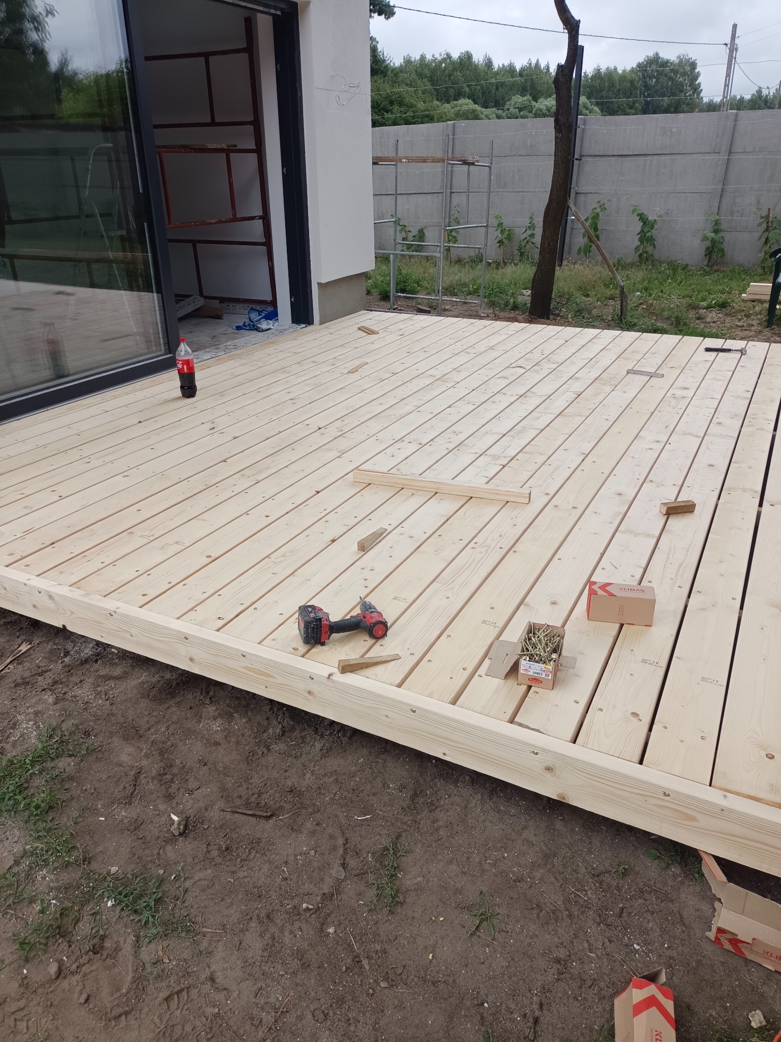 Budowa drewnianego tarasu: deski ułożone na ramie, wkrętarka, pudełko z wkrętami Klimas, butelka coli, widok na ogród i częściowo wykończony budynek z dużym oknem.