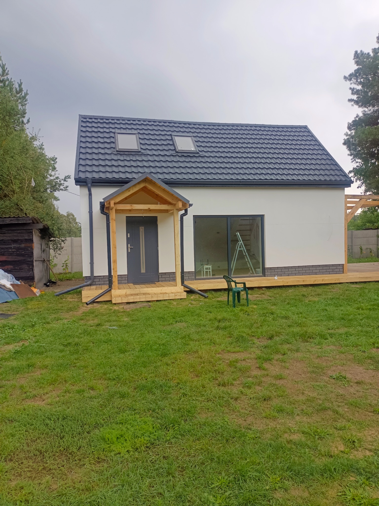Nowoczesny dom jednorodzinny z szarą dachówką, drewnianym podestem i zadaszeniem nad wejściem, widok z zewnątrz na trawnik w pochmurny dzień.
