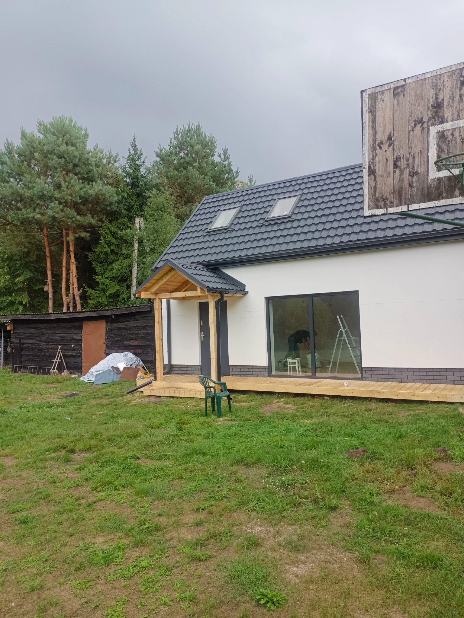 Nowoczesny dom z szarym dachem, drewnianym tarasem i zadaszeniem nad wejściem, widoczny kosz do koszykówki na ścianie, wnętrze w trakcie malowania widoczne przez okno.