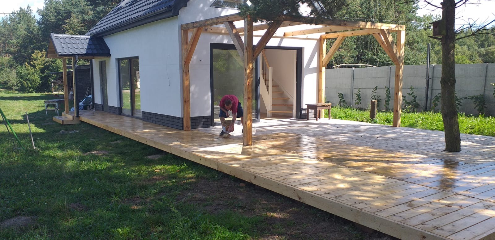 Pracownik impregnuje drewniany taras przy domu z białymi ścianami i drewnianą pergolą, widoczny fragment ogrodu i betonowe ogrodzenie.