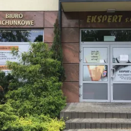 Fasada budynku z napisem 'Biuro Rachunkowe' i 'Ekspert s.c.', wejście z szarymi drzwiami, przed budynkiem ozdobne krzewy i schody z kostki.