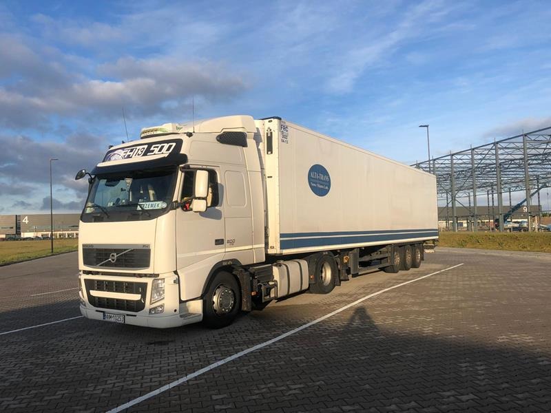 Biała ciężarówka Volvo FH 500 z naczepą chłodniczą Alta-Trans parkuje na placu z kostki brukowej w słoneczny dzień, widoczna konstrukcja hali w tle.