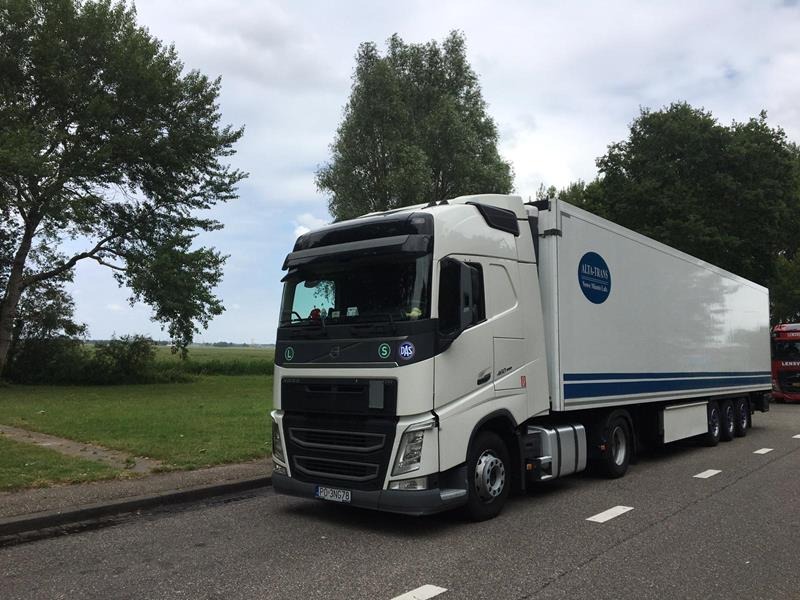 Biała ciężarówka Volvo z naczepą, z logo firmy Alta-Trans, zaparkowana na szosie w pobliżu zielonego pola i drzew, z widoczną tablicą rejestracyjną.