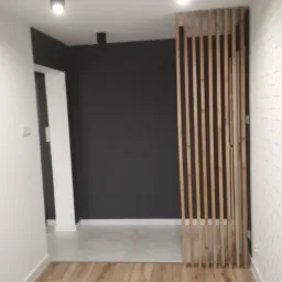 Nowoczesny przedpokój z drewnianą ażurową ścianką działową, podłoga z paneli, ściany w kolorze białym i grafitowym, sufit z oświetleniem punktowym.