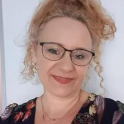 Portret kobiety w okularach z kręconymi, rudymi włosami, uśmiechającej się do kamery, w bluzce z motywem kwiatowym i dwoma delikatnymi naszyjnikami