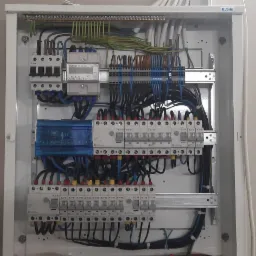 Otwarta skrzynka elektryczna z zainstalowanymi bezpiecznikami, licznikiem i okablowaniem, widoczna na tle ściany z fragmentem kamiennej konstrukcji.