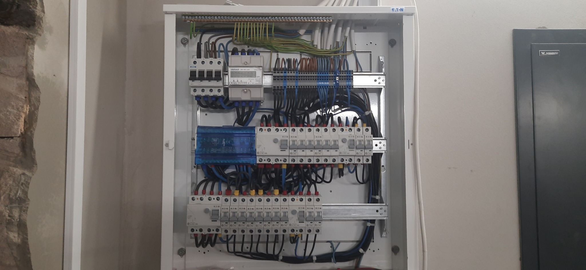 Otwarta skrzynka elektryczna z zainstalowanymi bezpiecznikami, licznikiem i okablowaniem, widoczna na tle ściany z fragmentem kamiennej konstrukcji.