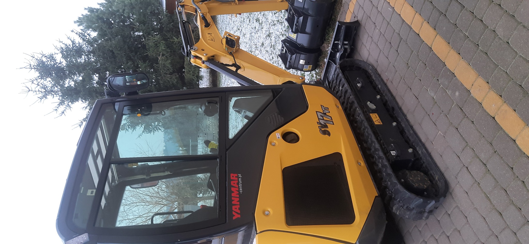 Żółto-czarna mini koparka Yanmar SV17VT na kostce brukowej, widoczne gąsienice i ramię koparki, w tle drzewo iglaste.