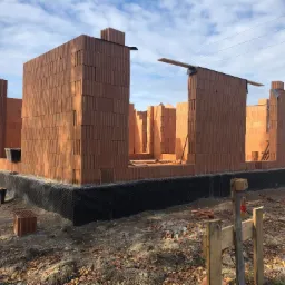 Wznoszone ściany parteru domu z czerwonej ceramiki poryzowanej, widoczna hydroizolacja fundamentów, drewniane stemple i fragment białego samochodu w tle.