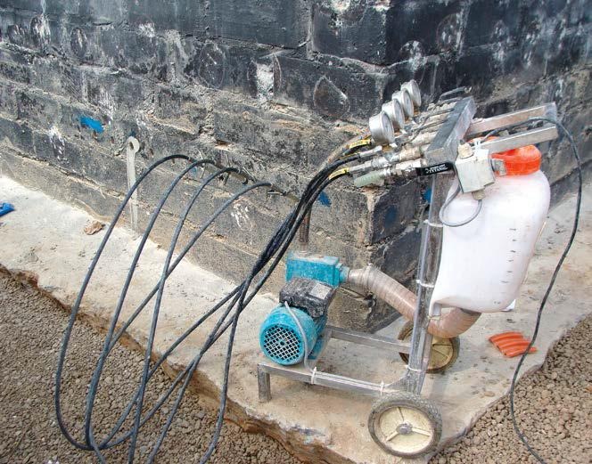 Sprzęt do iniekcji w zawilgoconej ścianie z cegły, widoczne węże, manometry i zbiornik, na zewnątrz budynku.