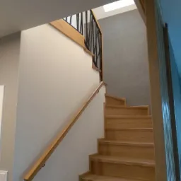 Jasne, drewniane schody z czarną, metalową balustradą, oświetlone światłem z okna dachowego, widok z dołu.