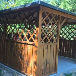 Drewniana pergola z kratownicą i niskimi panelami, ustawiona w ogrodzie, z widocznymi drzewami w tle.