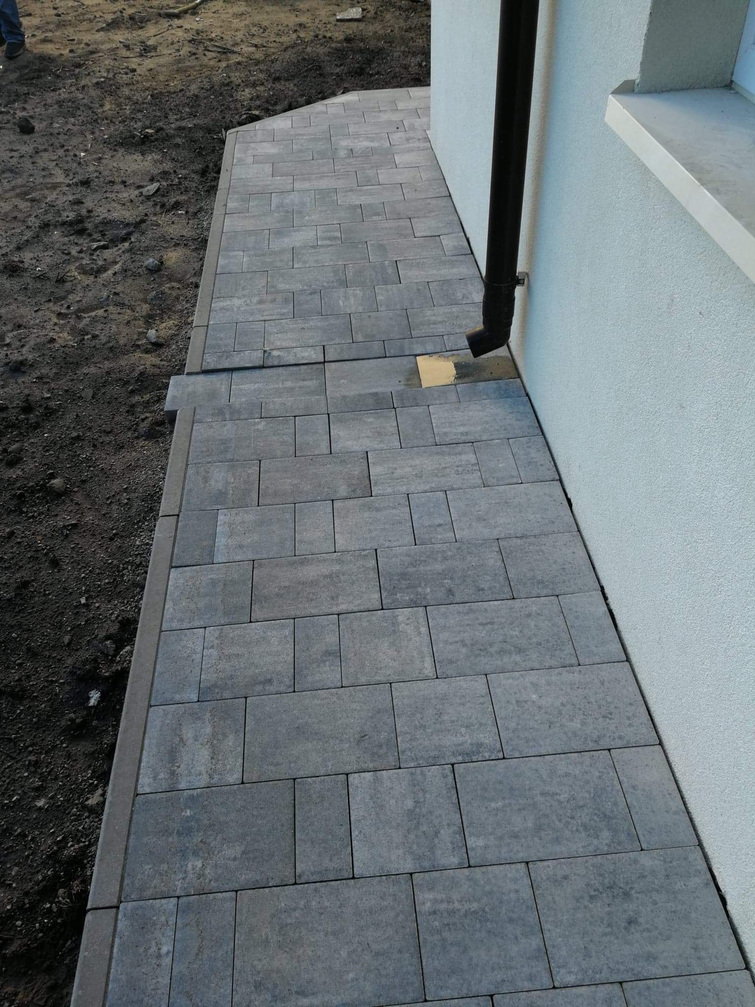 Szara kostka brukowa ułożona wzdłuż ściany budynku z rurą spustową, widoczny fragment nieukończonej nawierzchni z ziemią.