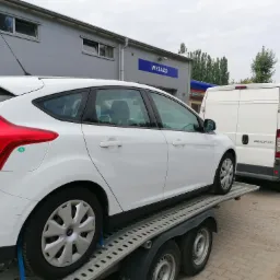 Biały Ford Focus zabezpieczony pasami na lawecie transportowej, za nim biały bus marki Ducato na tle budynku z napisem 'WYJAZD'.