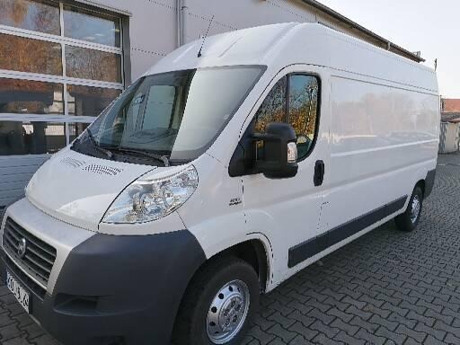 Biały samochód dostawczy Fiat Ducato z widoczną tablicą rejestracyjną, zaparkowany na brukowanej nawierzchni przed budynkiem z szarą bramą garażową.
