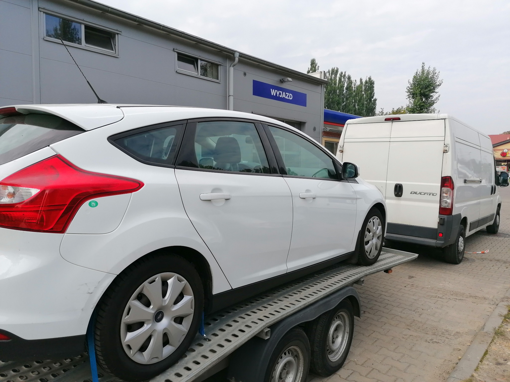 Biały Ford Focus zabezpieczony pasami na lawecie transportowej, za nim biały bus marki Ducato na tle budynku z napisem 'WYJAZD'.