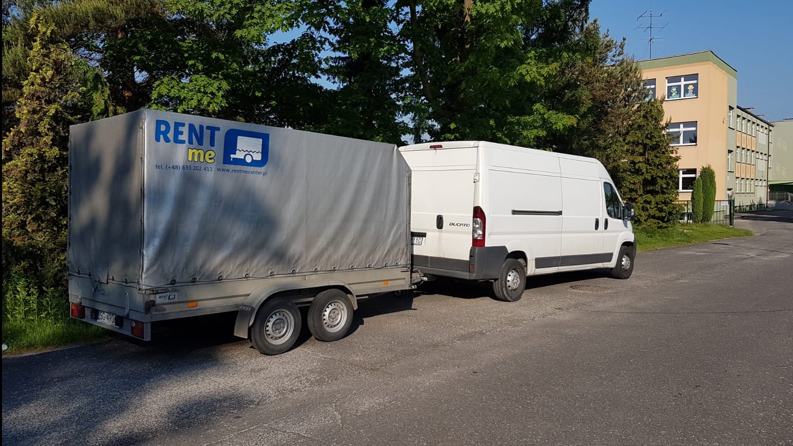 Biały bus z przyczepą z plandeką z logo firmy RENT ME na tle zieleni i budynku w Rudzie Śląskiej.