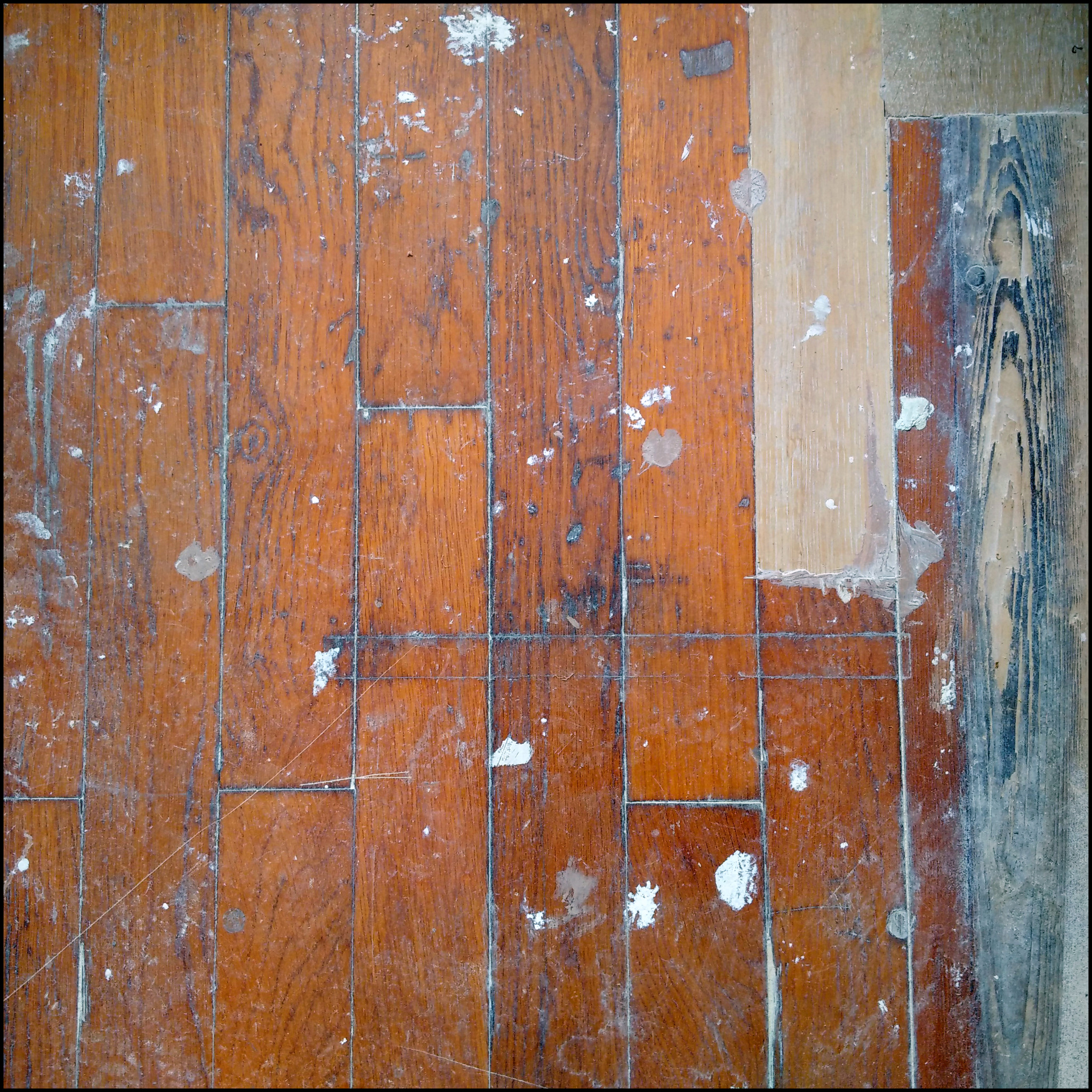 Fragment drewnianej podłogi parkietowej z widocznymi śladami farby i zarysowaniami, częściowo odnowiona z jedną deską w surowym drewnie i fragmentem z ciemnym, postarzanym wykończeniem.