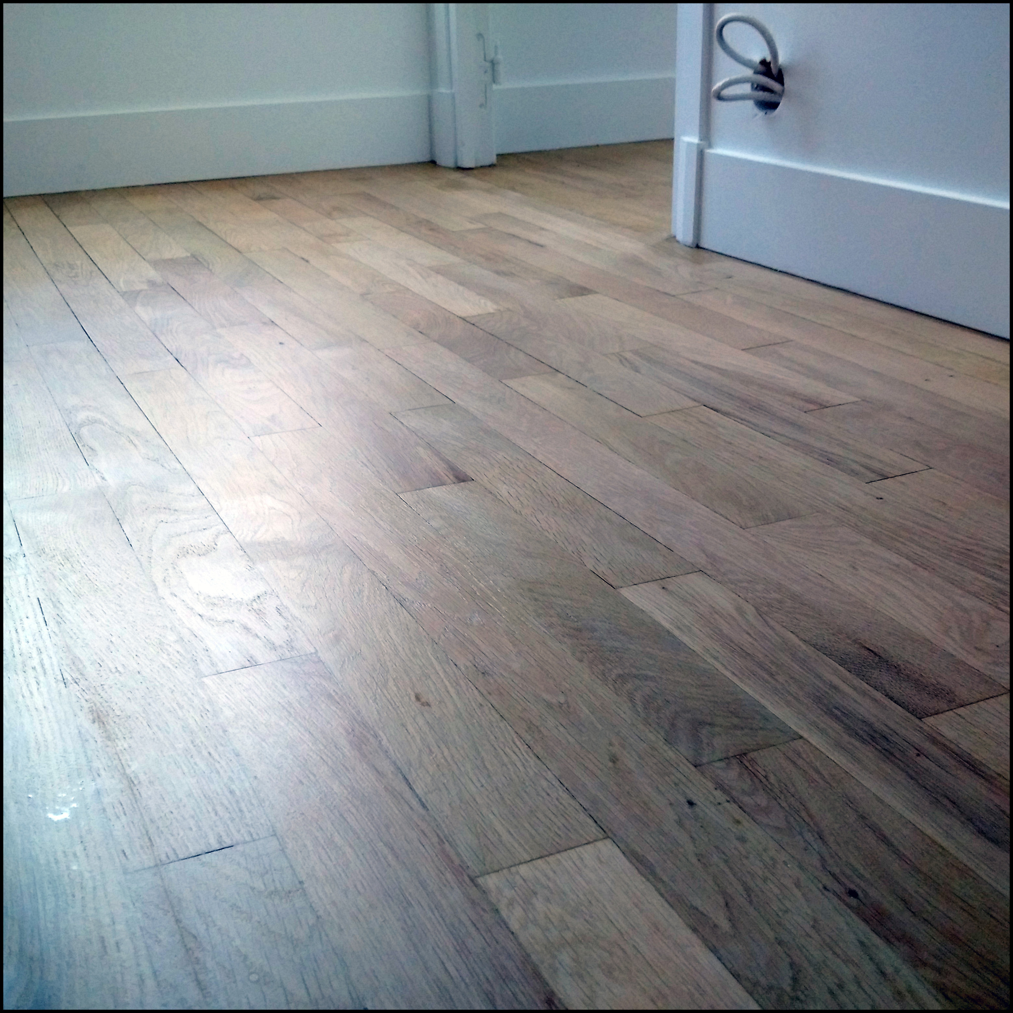 Wycyklinowana podłoga drewniana z widocznymi śladami użytkowania i połyskiem, białe listwy przypodłogowe i fragment otwartych drzwi.