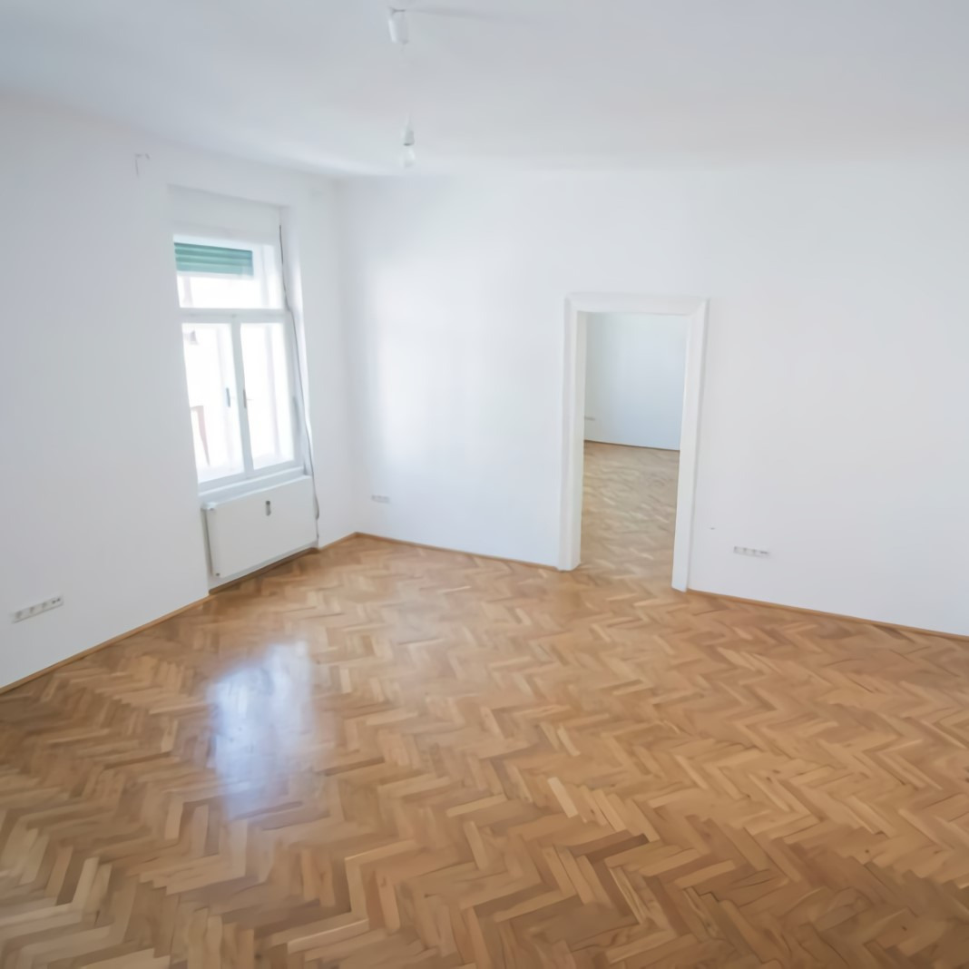 Pusty, jasny pokój z parkietem ułożonym w jodełkę, po renowacji, z widocznymi śladami po cyklinowaniu.