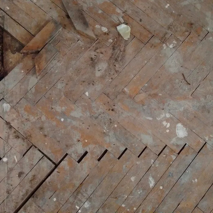 Drewniana podłoga ułożona w jodełkę, widoczne uszkodzenia, ubytki i ślady farby przed renowacją.