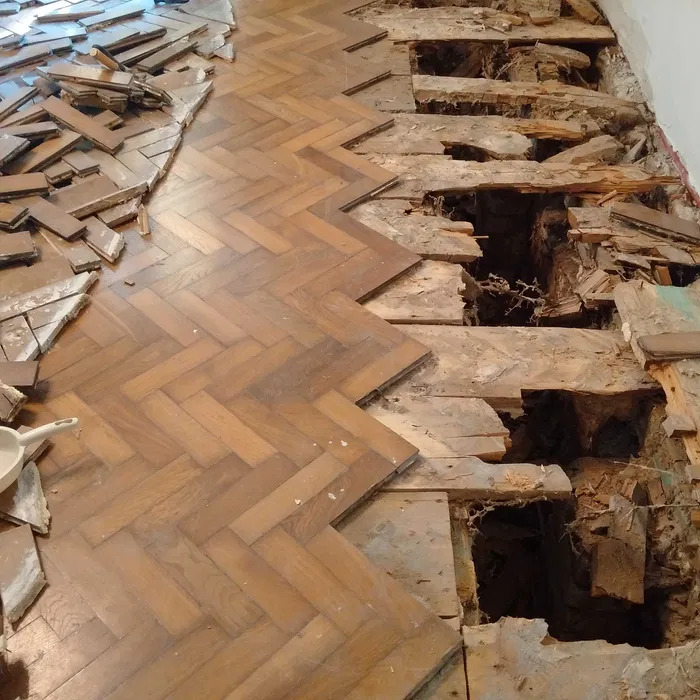 Fragment podłogi z parkietem ułożonym w jodełkę, częściowo odnowiony, z widocznymi ubytkami i uszkodzeniami po prawej stronie oraz stertą parkietu do ułożenia po lewej.