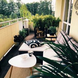 Dan72 - Przytulny balkon z drewnianą podłogą, meblami rattanowymi, poduszkami w geometryczne wzory i licznymi roślinami w donicach, w tle widok na zieleń drzew.