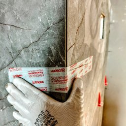 Dan72 - Układanie płytek imitujących marmur, zabezpieczonych taśmą tynkarską Dolphin, widoczne czerwone kliny poziomujące i dłoń w rękawicy roboczej M-Glove.