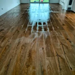 Świeżo polakierowana, błyszcząca drewniana podłoga ułożona w pustym pokoju z dużym oknem balkonowym i niedokończonymi ścianami.